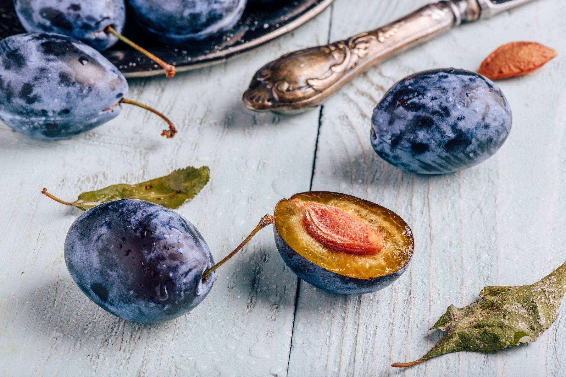 Ripe blue-black plums, one halved to show its amber flesh, on weathered white wood